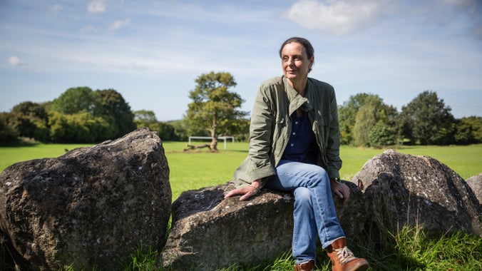 A women wearing a green jacket and jeans sits on a row of rocks in a park